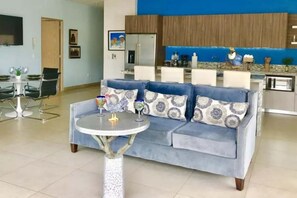 Un salon avec un canapé bleu, une table basse ronde et une cuisine en arrière-plan.