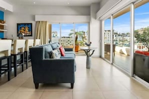Un salon moderne avec un canapé, un coin repas avec des chaises, une grande fenêtre donnant sur des immeubles et une petite table avec une plante.