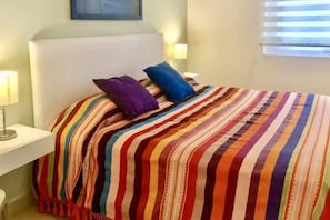 A bed with a striped bedspread, two pillows, a bedside table with a lamp, and a window with blinds. - Zenith Penthouse 702 | Rooftop Pool & Ocean Views (Puerto Vallarta)