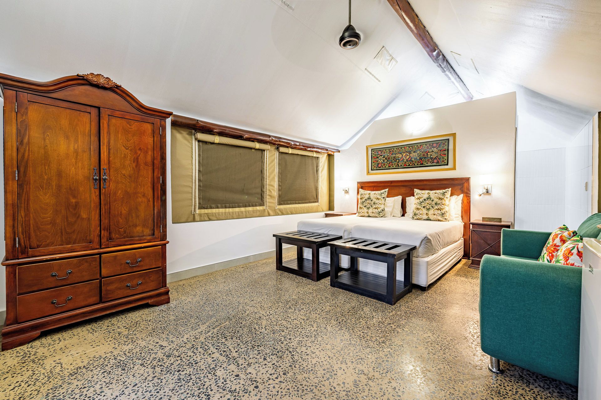 A bedroom with a large wooden wardrobe, a bed with floral pillows, a green armchair, and a small table.