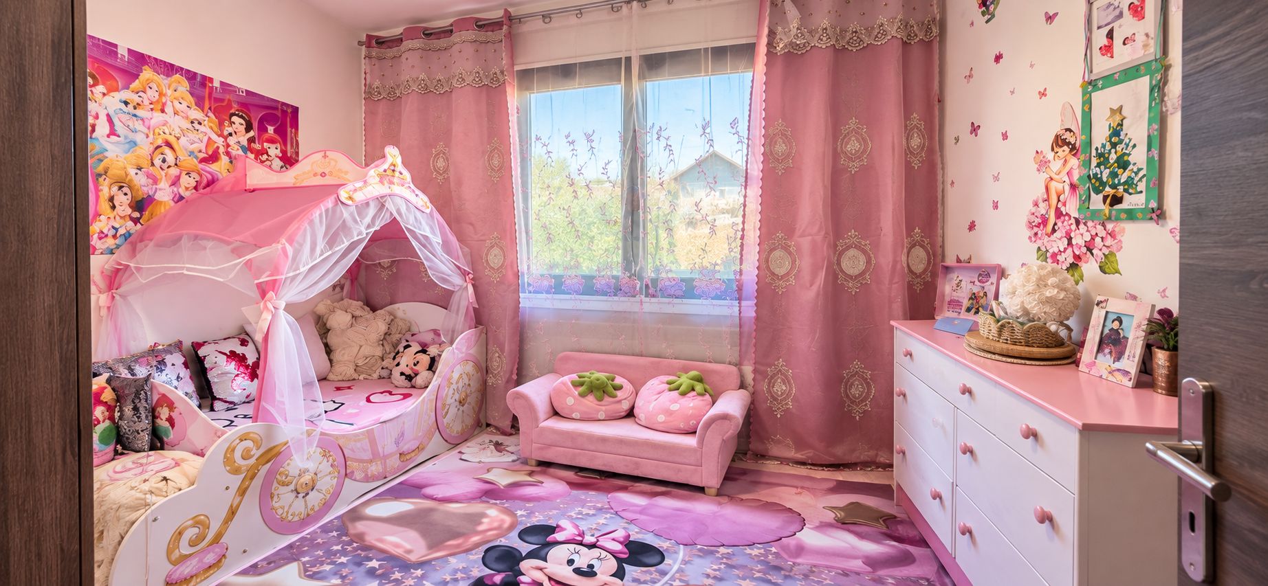 Een kamer voor kinderen, ingericht in roze tinten, met een bed, een roze bank, een dressoir en een raam met gordijnen.