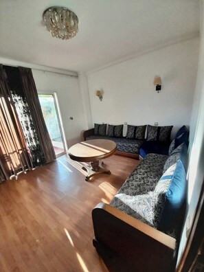 Ein großzügiger Wohnraum mit Holzboden, einem runden Couchtisch, einer Couch mit gemusterten Kissen und einem großen Fenster mit Vorhängen.