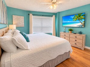 Un dormitorio con cama, mesita de noche, cómoda, ventilador de techo, ventana con persianas y un televisor de pared mostrando una escena de playa.