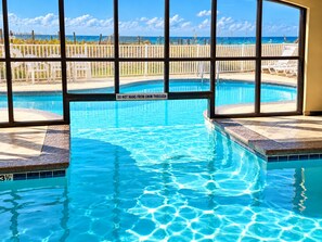 Une piscine intérieure à l’eau d’un bleu limpide, un panneau d’avertissement et une vue sur la plage grâce à de grandes fenêtres.