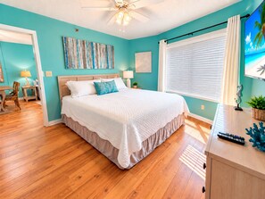 Une chambre à coucher avec un lit, un plancher en bois, un ventilateur de plafond, une fenêtre avec des stores et un mur orné d’un tableau.