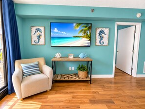 Una habitación con una pared verde azulado, un televisor sobre una mesa de madera, un sillón color beige con un cojín a rayas y una puerta que da a otra habitación.