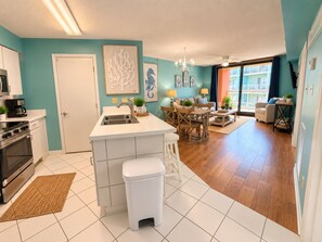 A modern kitchen with a central island, a dining area with wooden chairs, and a view of the outside balcony. - Cute 2 Bed 2 Bath at Harbor Place - Fall Specials! (Orange Beach)