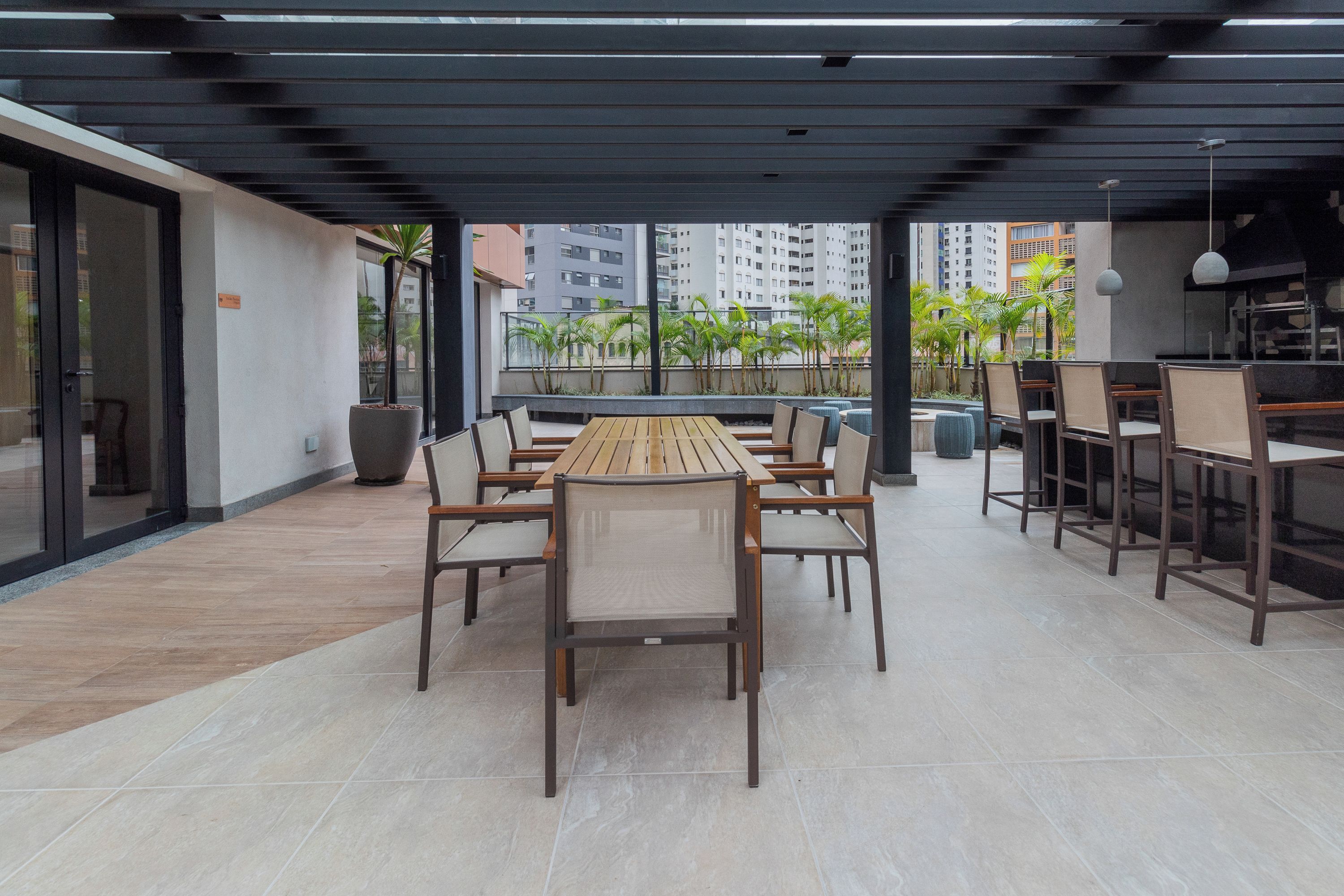 An outdoor dining area with a long wooden table and chairs, surrounded by a covered patio with a view of palm trees and buildings.