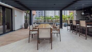 An outdoor dining area with a long wooden table and chairs, surrounded by a covered patio with a view of palm trees and buildings.