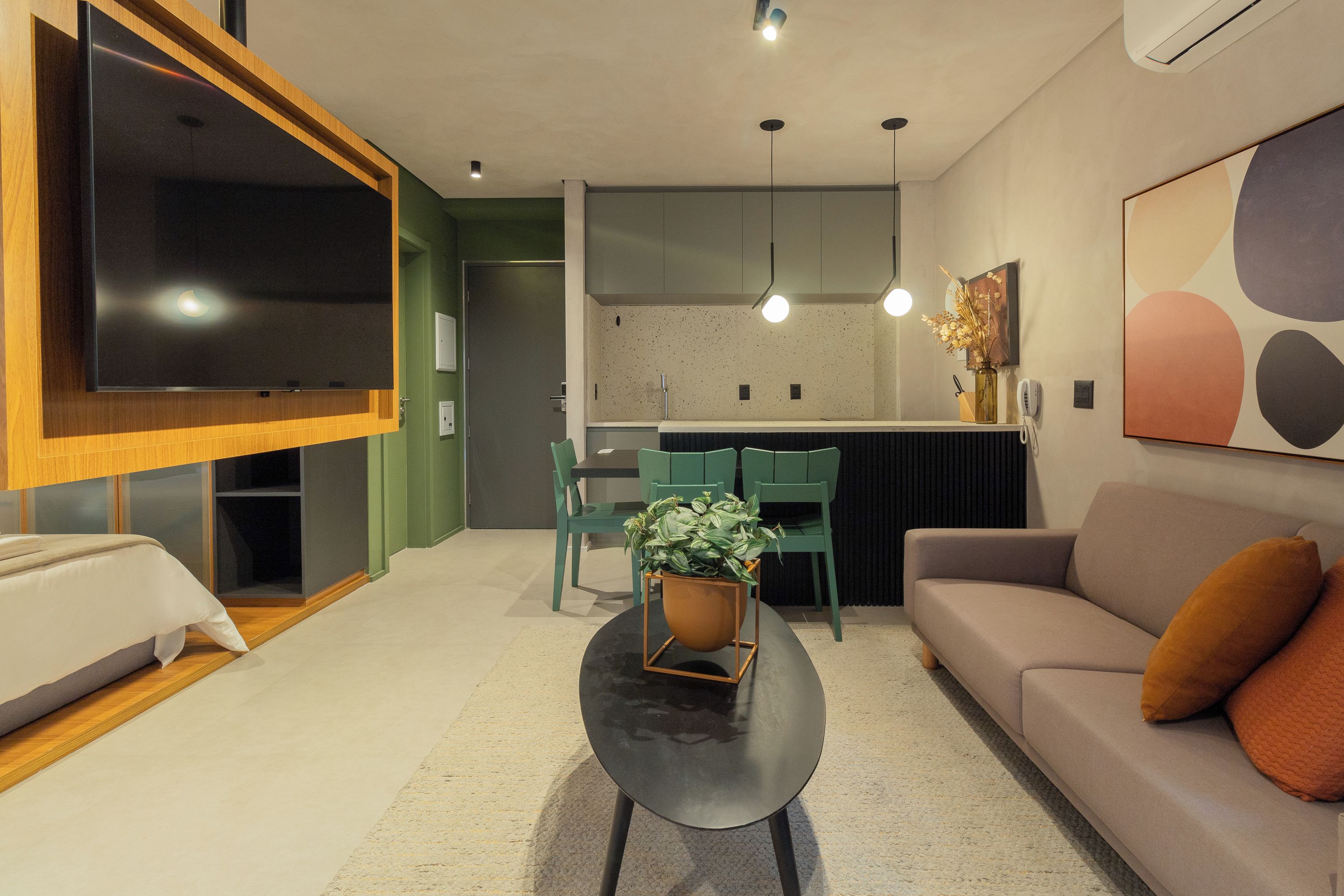 A modern living room with a sofa, a coffee table with a plant, a TV mounted on a wooden panel, and a kitchen area with green chairs.