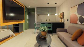 A modern living room with a sofa, a coffee table with a plant, a TV mounted on a wooden panel, and a kitchen area with green chairs.