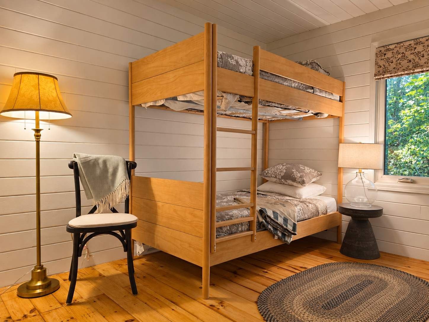 Ein Stockbett mit einer Leiter, ein Stuhl, eine Lampe und ein runder Teppich in einem Zimmer mit Holzboden und einem Fenster mit Blick auf Grünflächen.