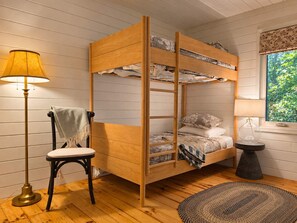 Een stapelbed met een ladder, een stoel, een lamp en een rond vloerkleed in een kamer met houten vloer en een raam met uitzicht op groen.