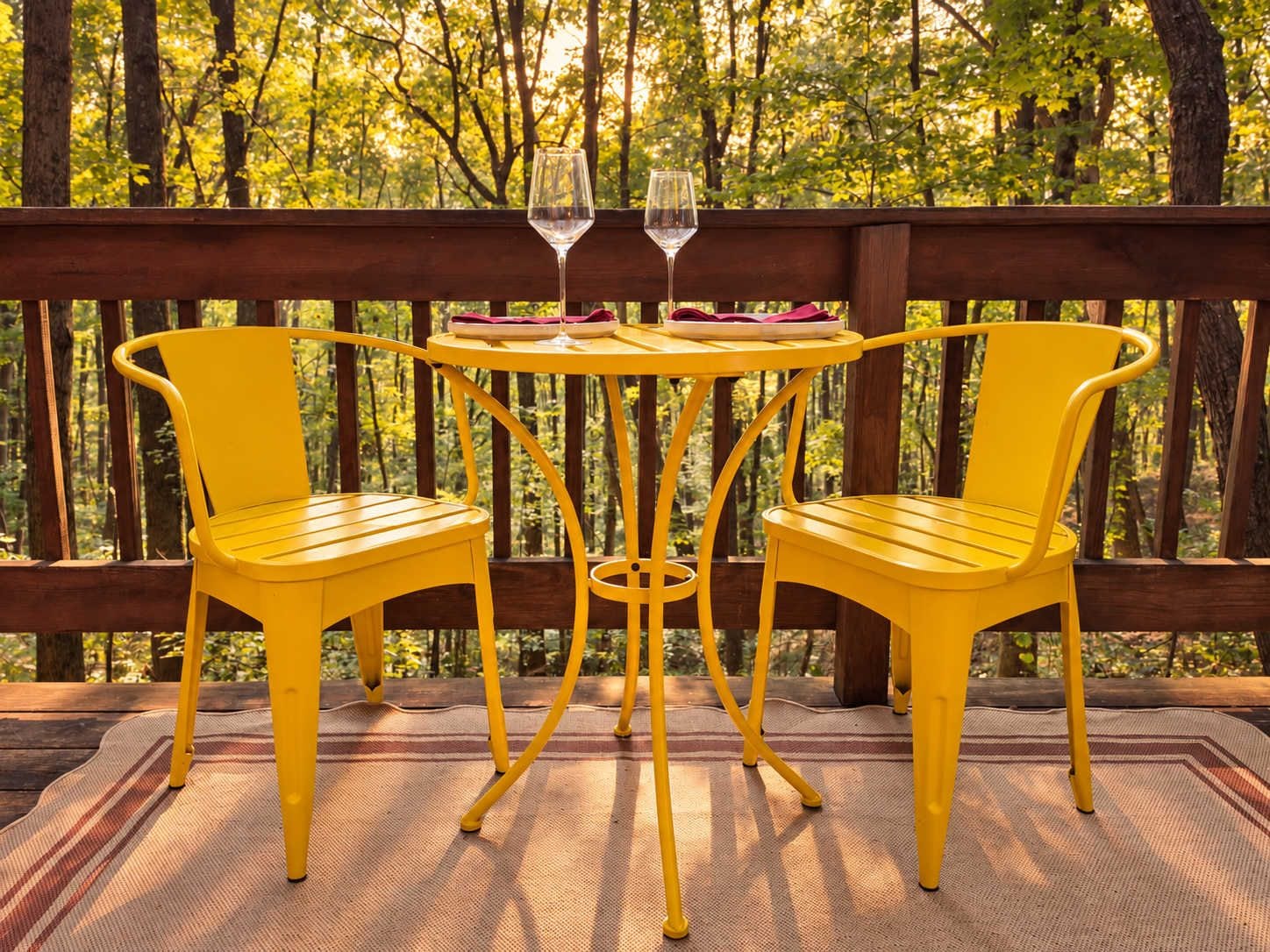 Een houten terras met twee gele stoelen en een kleine ronde tafel, waarop een glas wijn staat.