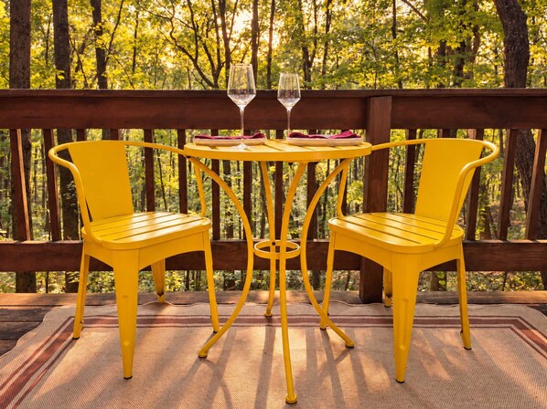 Une terrasse en bois avec deux chaises jaunes et une petite table ronde, sur laquelle se trouve un verre de vin.