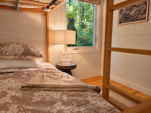 Une chambre à coucher avec un lit, une table de chevet, une lampe, une fenêtre donnant sur des arbres et un tableau encadré au mur.