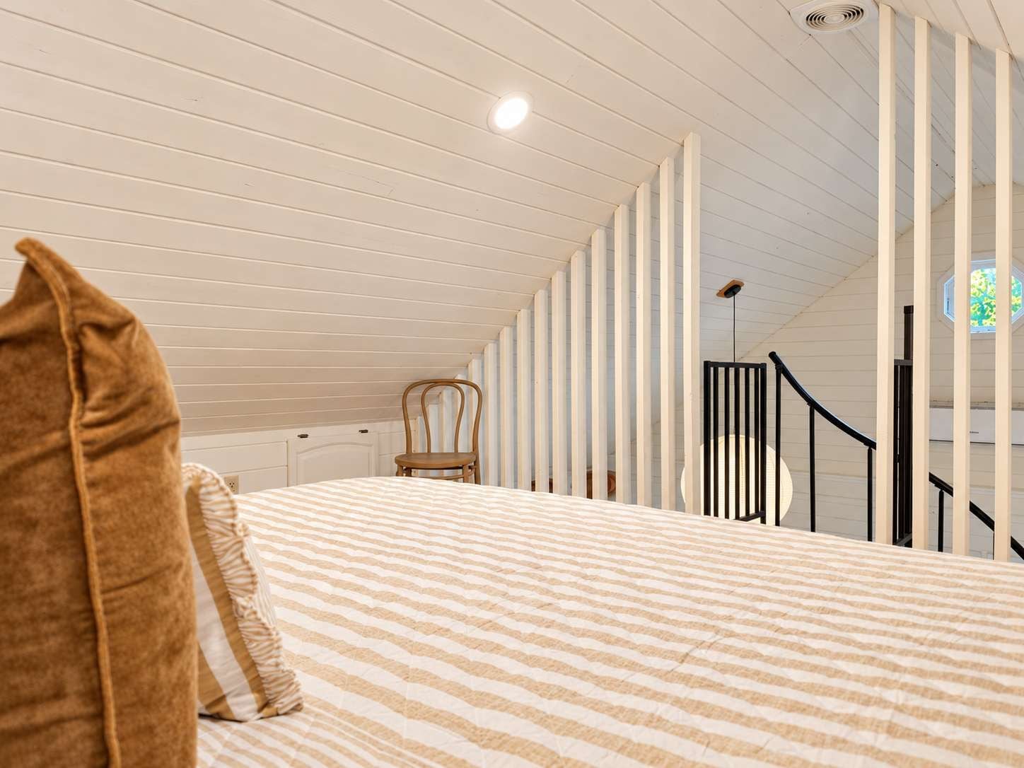 Un dormitorio con techo inclinado, una cama con ropa de cama a rayas, una silla y una ventana con vistas.