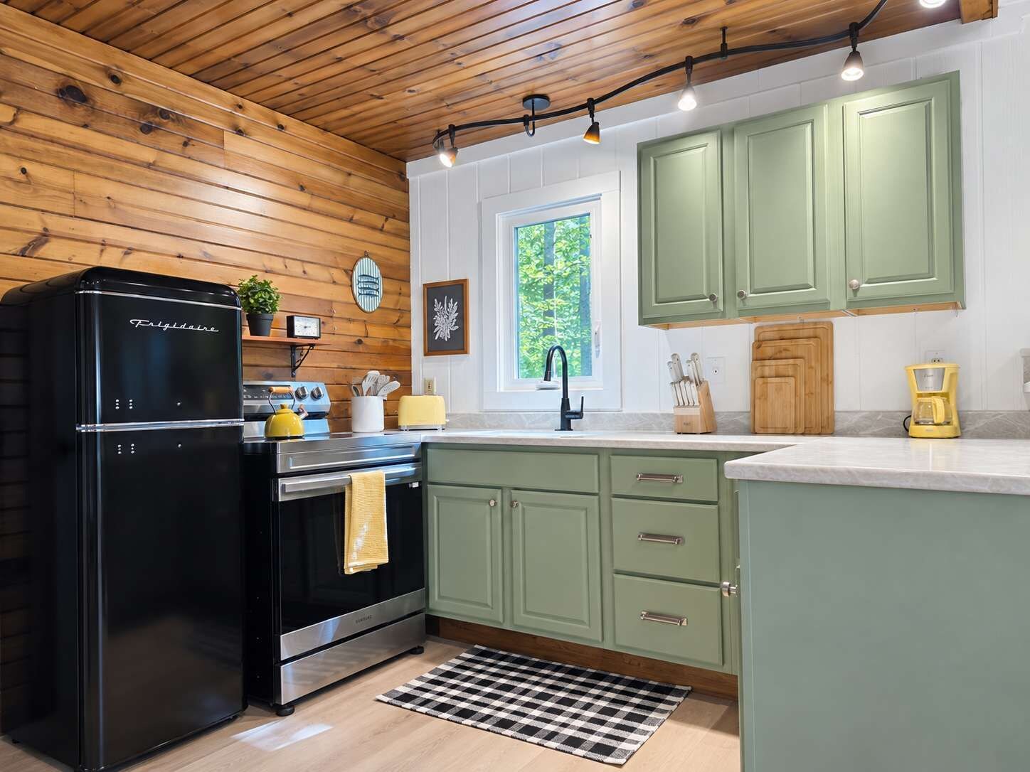 A kitchen with wooden ceiling, green cabinets, a black refrigerator, a stove, and a window with a view of greenery.