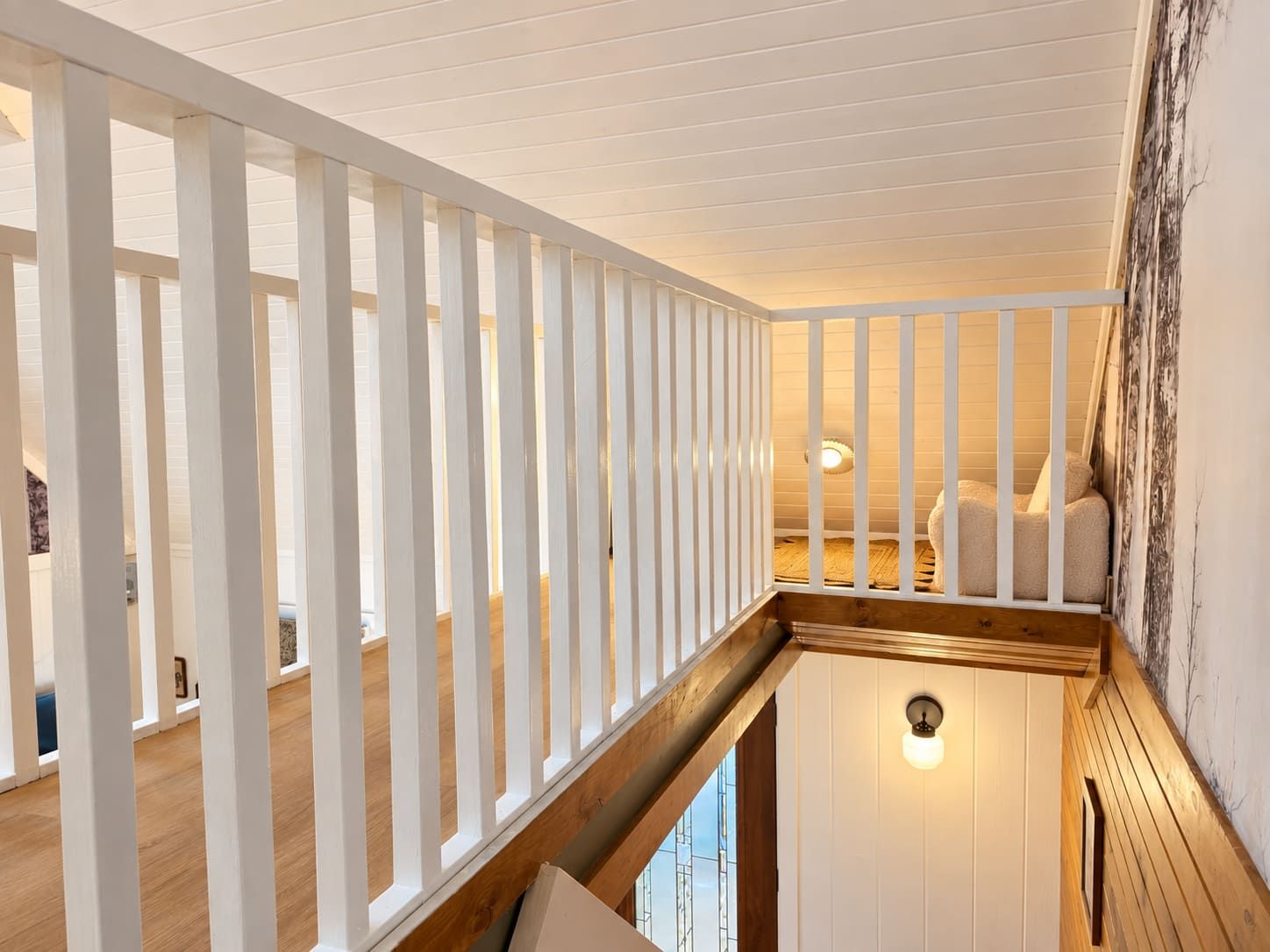 A room with a wooden ceiling, a white railing, a bed with pillows, and a wall-mounted light fixture.
