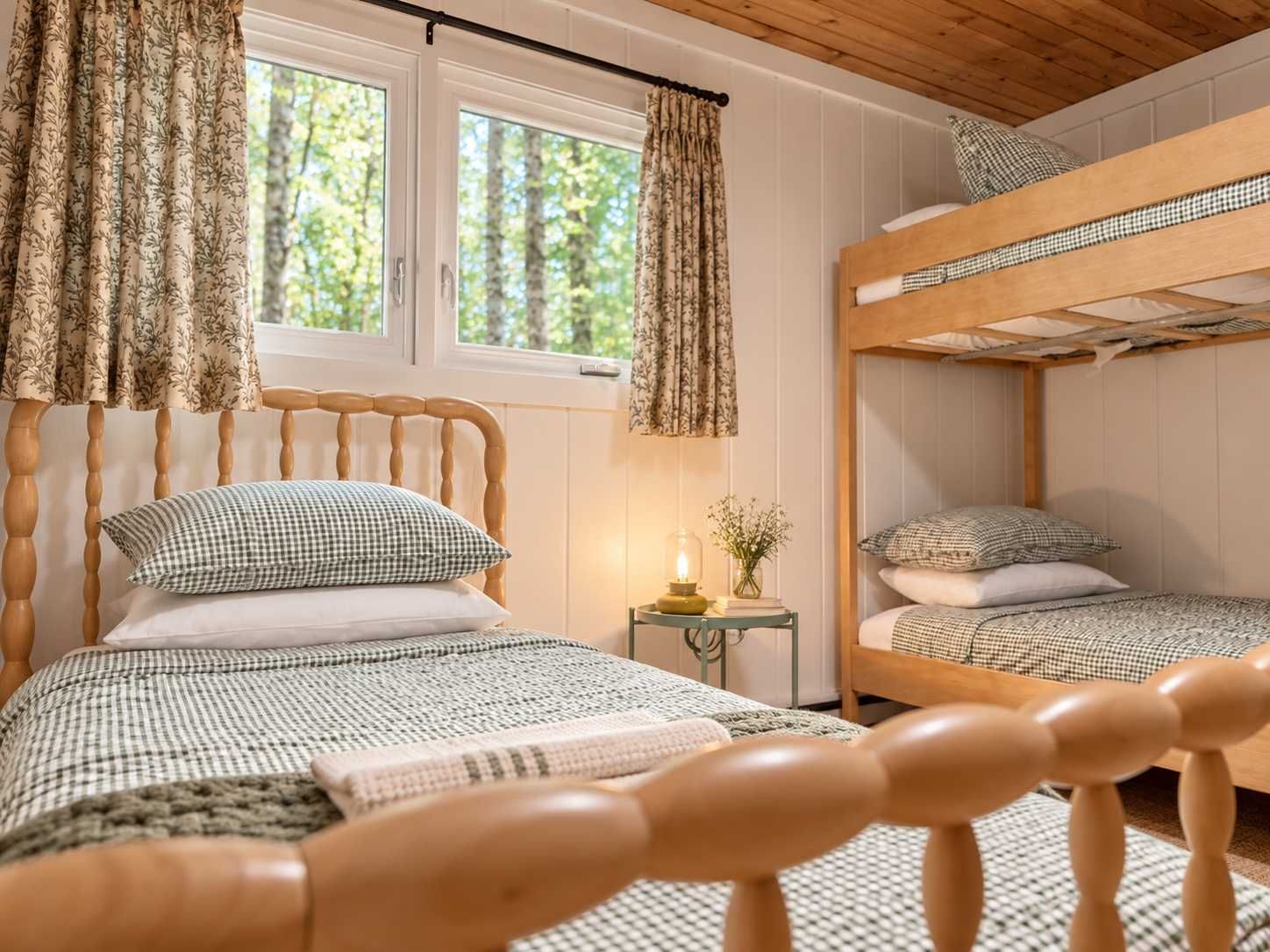 A room with a wooden bunk bed, a single bed with a checkered bedspread, and a window with curtains.