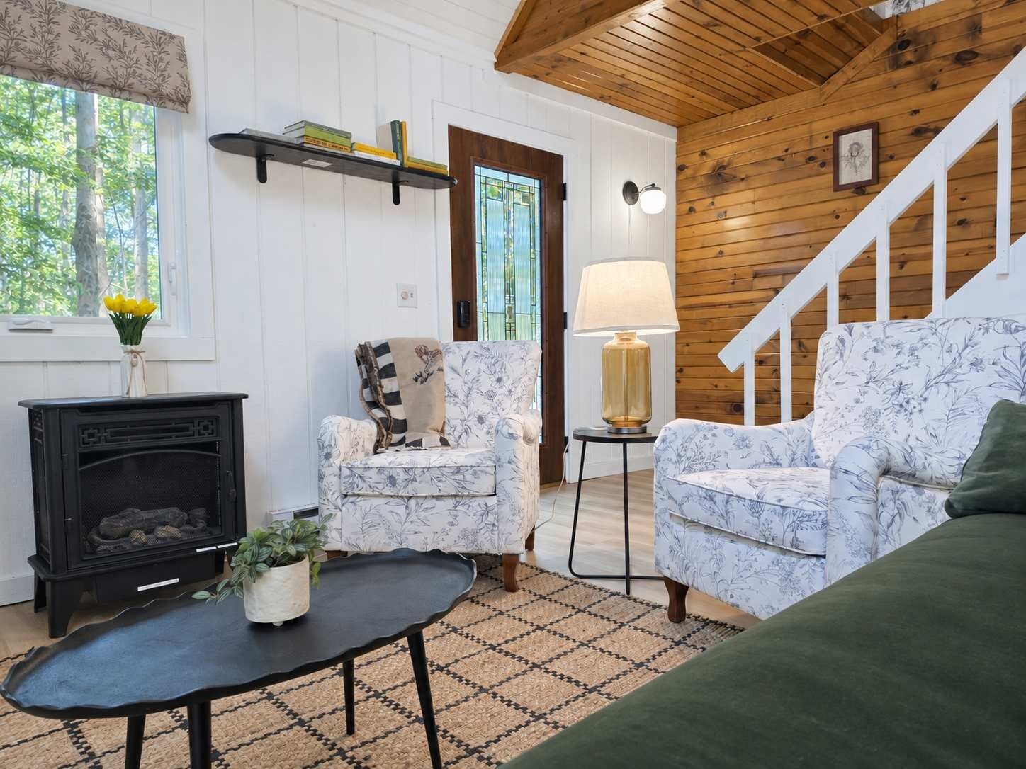 Una sala de estar acogedora con chimenea, dos sillones estampados, una mesita con una planta y una escalera que sube.