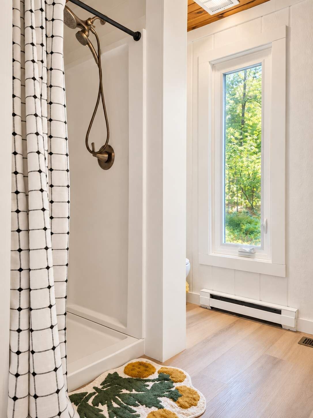 A bathroom with a shower, a window with a view of greenery, and a patterned rug on the floor.