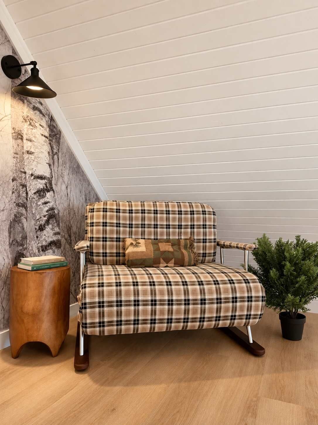 A plaid sofa with a cushion, a small wooden side table, a potted plant, and a wall-mounted lamp in a room with wooden flooring and a sloped ceiling.