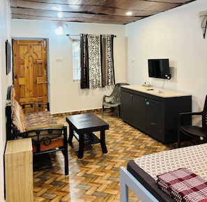 Quarto com porta de madeira, sofá, mesa de jantar, televisão e piso estampado.