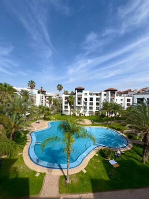 A resort with a large swimming pool surrounded by palm trees and a clear blue sky. - Comfortable , quiet , and beautiful apartment. (Agadir)