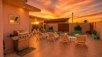 An outdoor patio with a grill, lounge chairs, and a pool under a sunset sky.