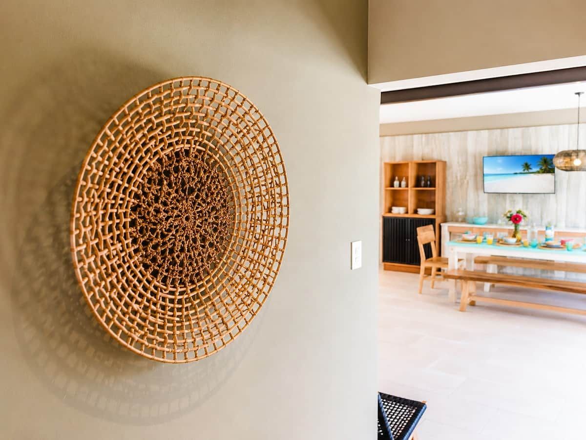 A modern interior with a circular woven light fixture on a wall, a dining area with a wooden table and chairs, and a television mounted on the wall.