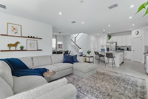 Ein modernes Wohnzimmer mit einem Ecksofa, einem Essbereich mit einer Kronleuchter und einer Küche mit weißen Schränken.