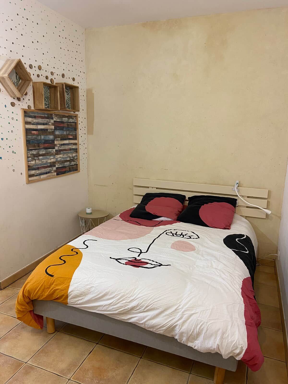 Ein Bett mit weißroter Steppdecke, ein kleiner Nachttisch mit Lampe sowie eine Wand mit dekorativer Holzvertäfelung und einem gerahmten Kunstwerk.
