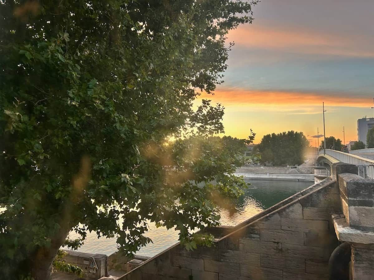 Ein Fluss mit einer Steinbrücke, Bäumen und einem Sonnenuntergang.