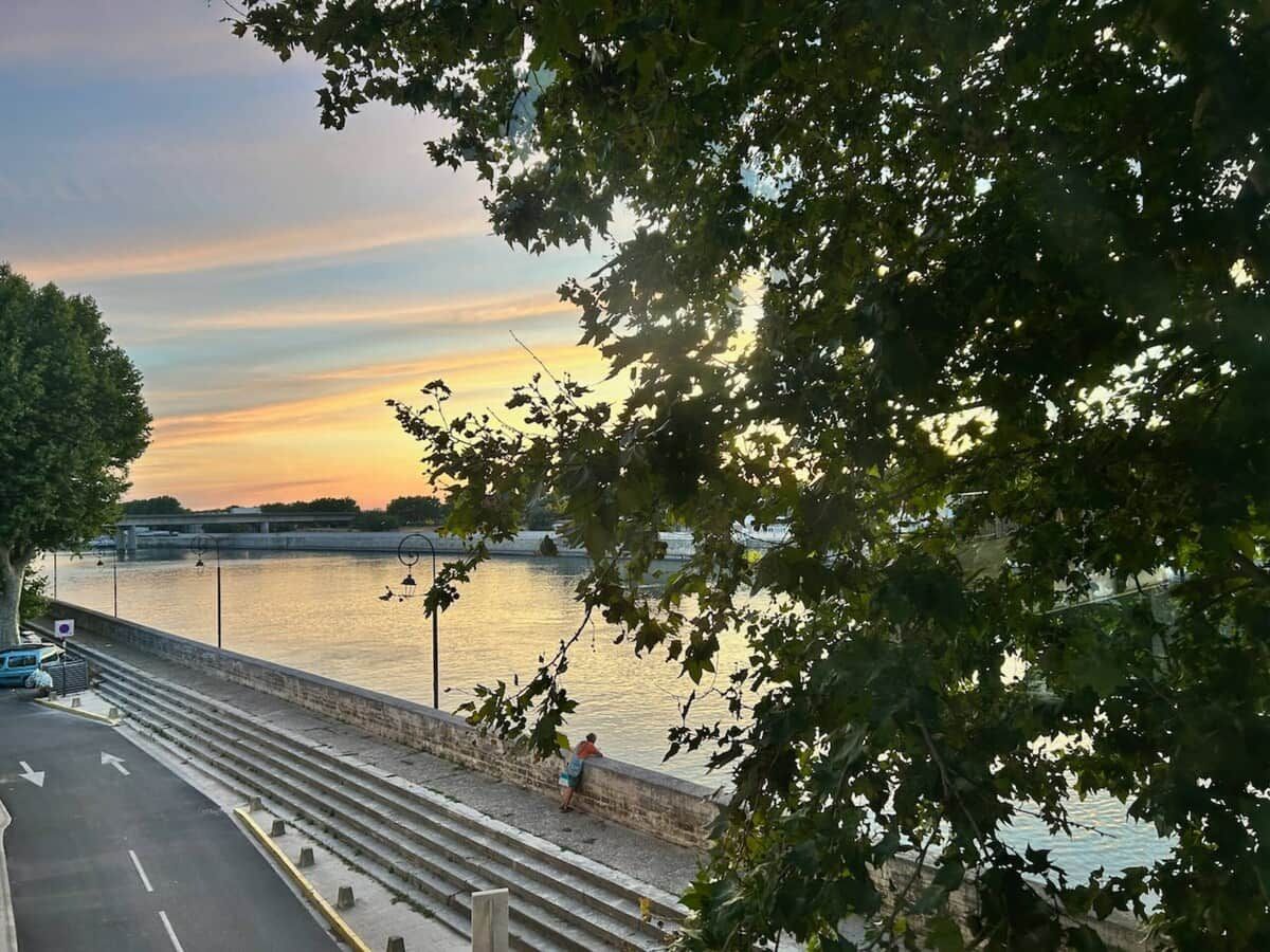 A riverside walkway with steps leading down to the water, a bridge in the background, and trees framing the scene.