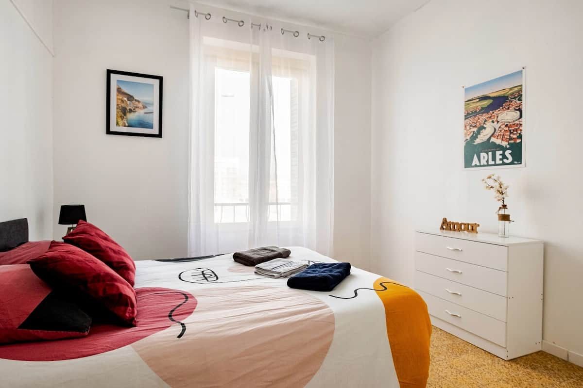 Una habitación de hotel con una cama, una mesita de noche, un aparador, un cartel en la pared y una ventana con cortinas de encaje.
