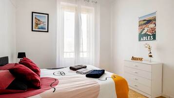 Una habitación de hotel con una cama, una mesita de noche, un aparador, un cartel en la pared y una ventana con cortinas de encaje.