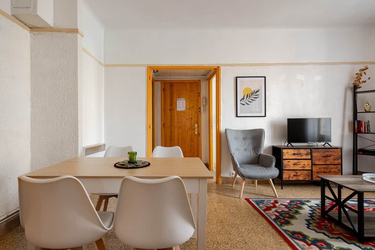 Un salon moderne avec une table à manger en bois, deux chaises blanches, un fauteuil gris, un téléviseur et une bibliothèque.