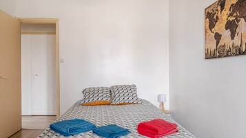 Un dormitorio con cama, almohadas y una manta. Hay una puerta y una pared con un cuadro.