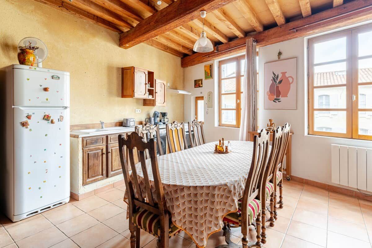 A dining area with a table covered by a patterned tablecloth, wooden chairs, a white refrigerator with magnets, wooden cabinets, and a window with a view.
