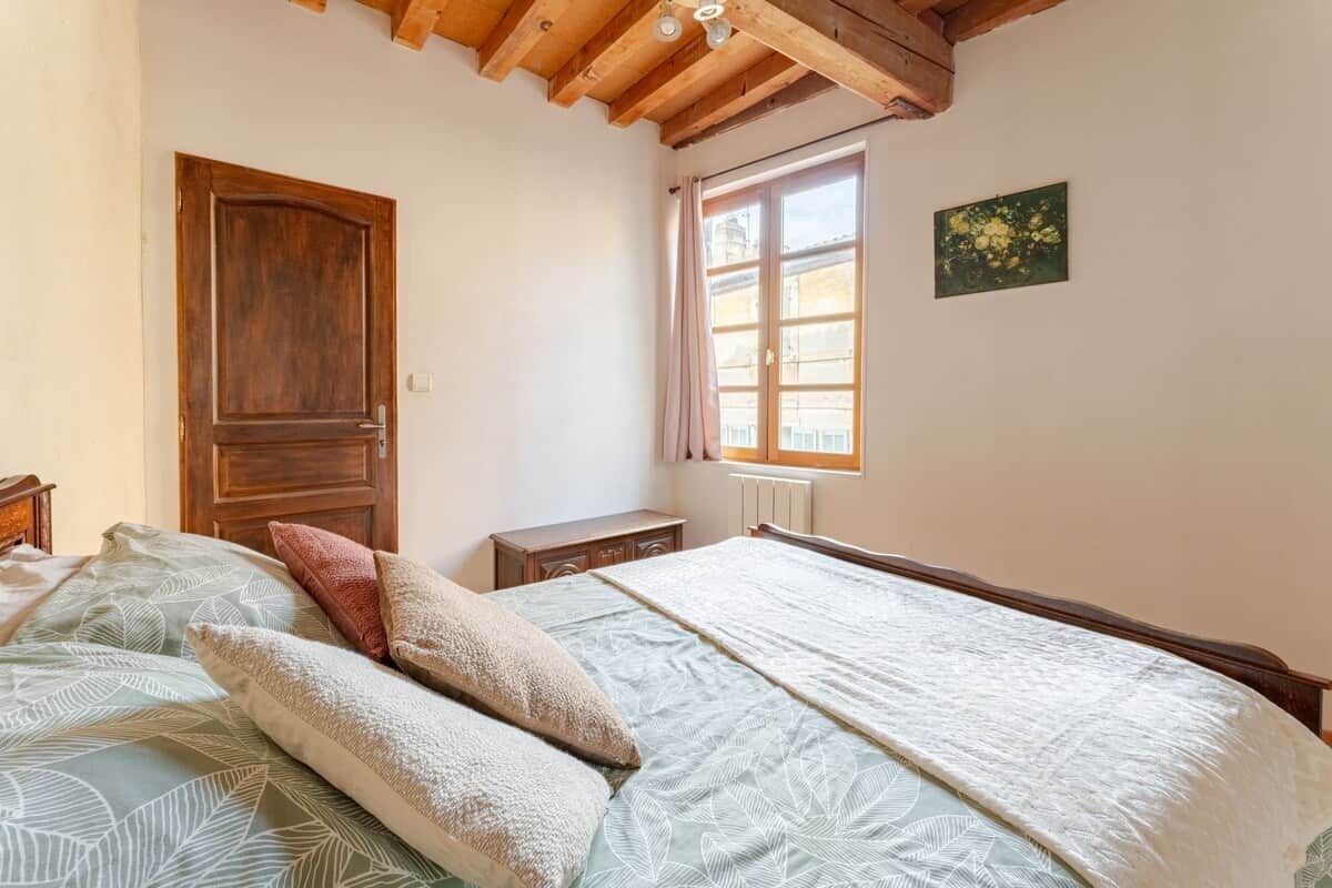 A bedroom with a wooden ceiling, a window with curtains, a bed with patterned bedding, and a small bedside table.