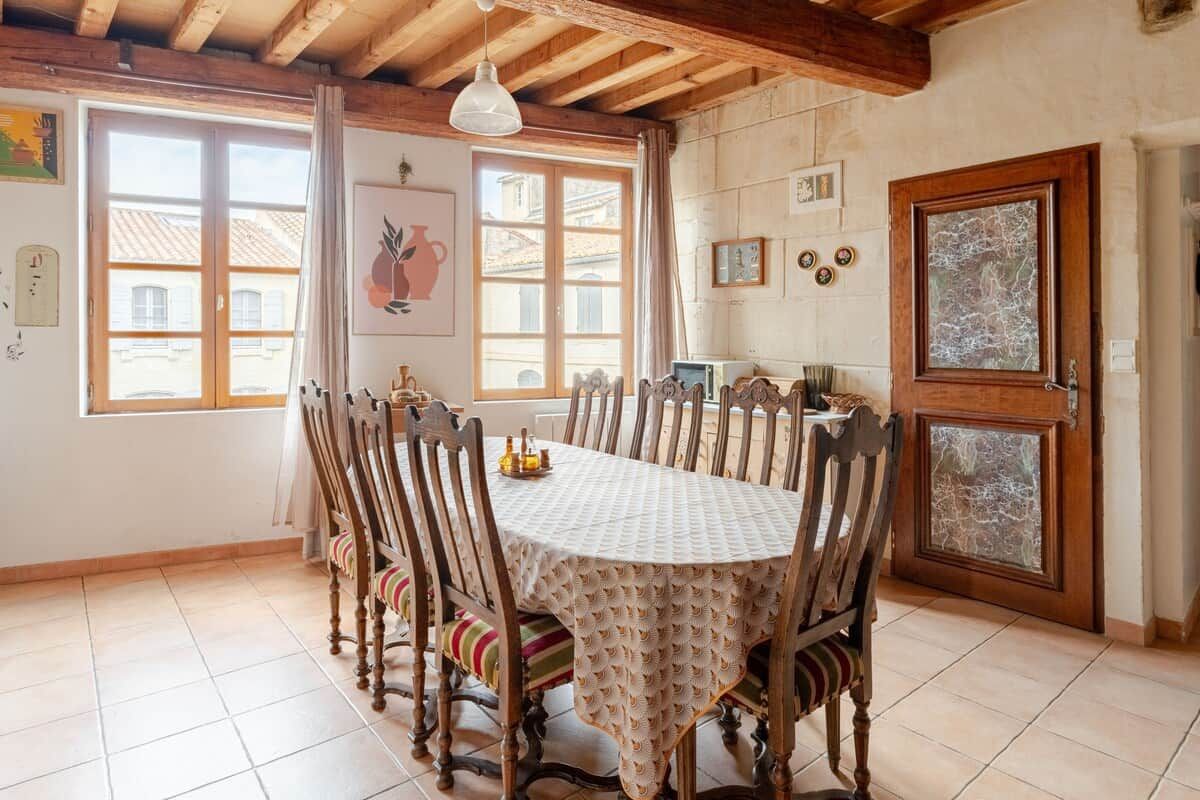 Un comedor con una larga mesa, sillas de madera y un mantel blanco. Hay ventanas con cortinas, una puerta con panel de vidrio y decoraciones en la pared.