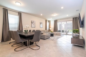 A modern living room with a dining area, a sofa, and a TV mounted on the wall. - Windsor Cay 7BR Themed Rooms, Pool & spa (Clermont)