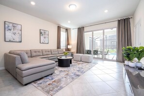 A modern living room with a sectional sofa, a round coffee table, and a view of a pool through large glass doors. - Windsor Cay 7BR Themed Rooms, Pool & spa (Clermont)