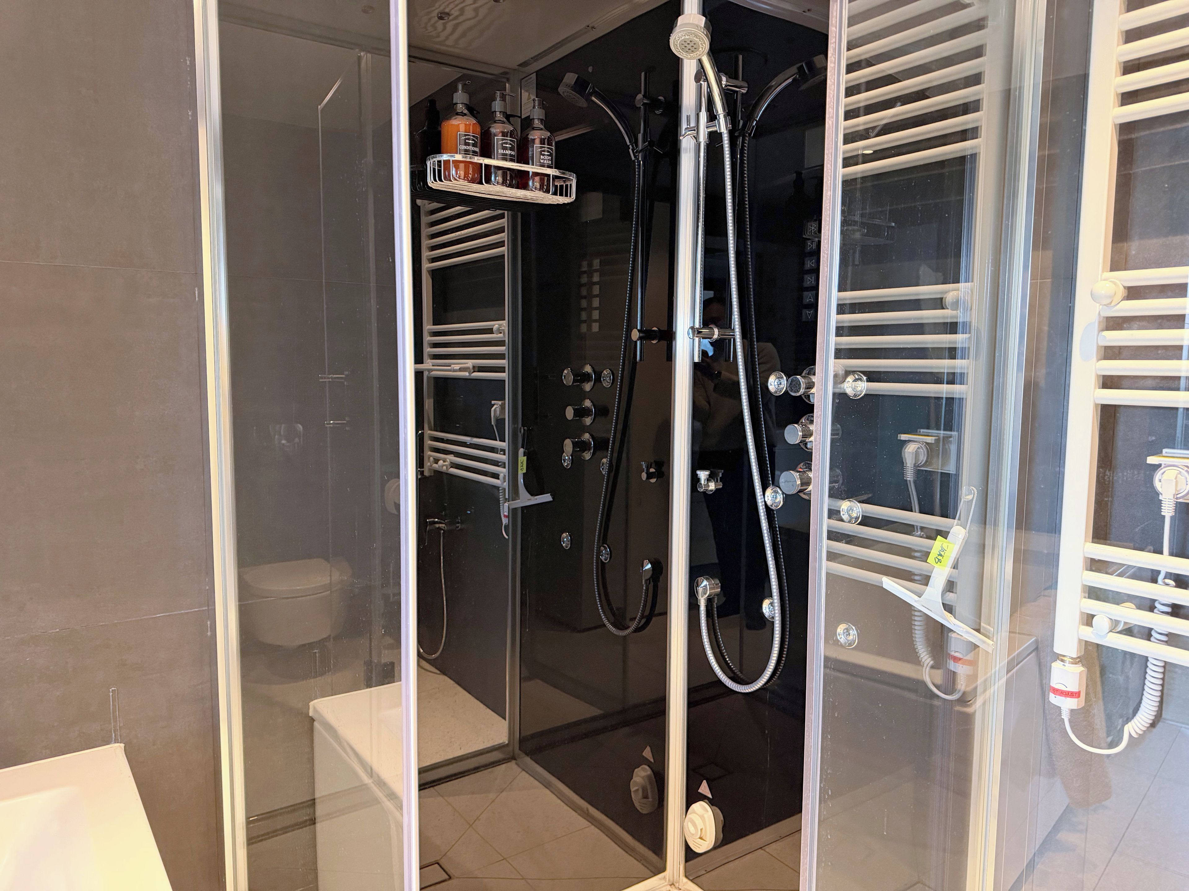 A glass-enclosed shower with multiple faucets and a shelf holding toiletries.