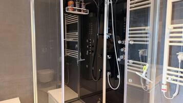A glass-enclosed shower with multiple faucets and a shelf holding toiletries.