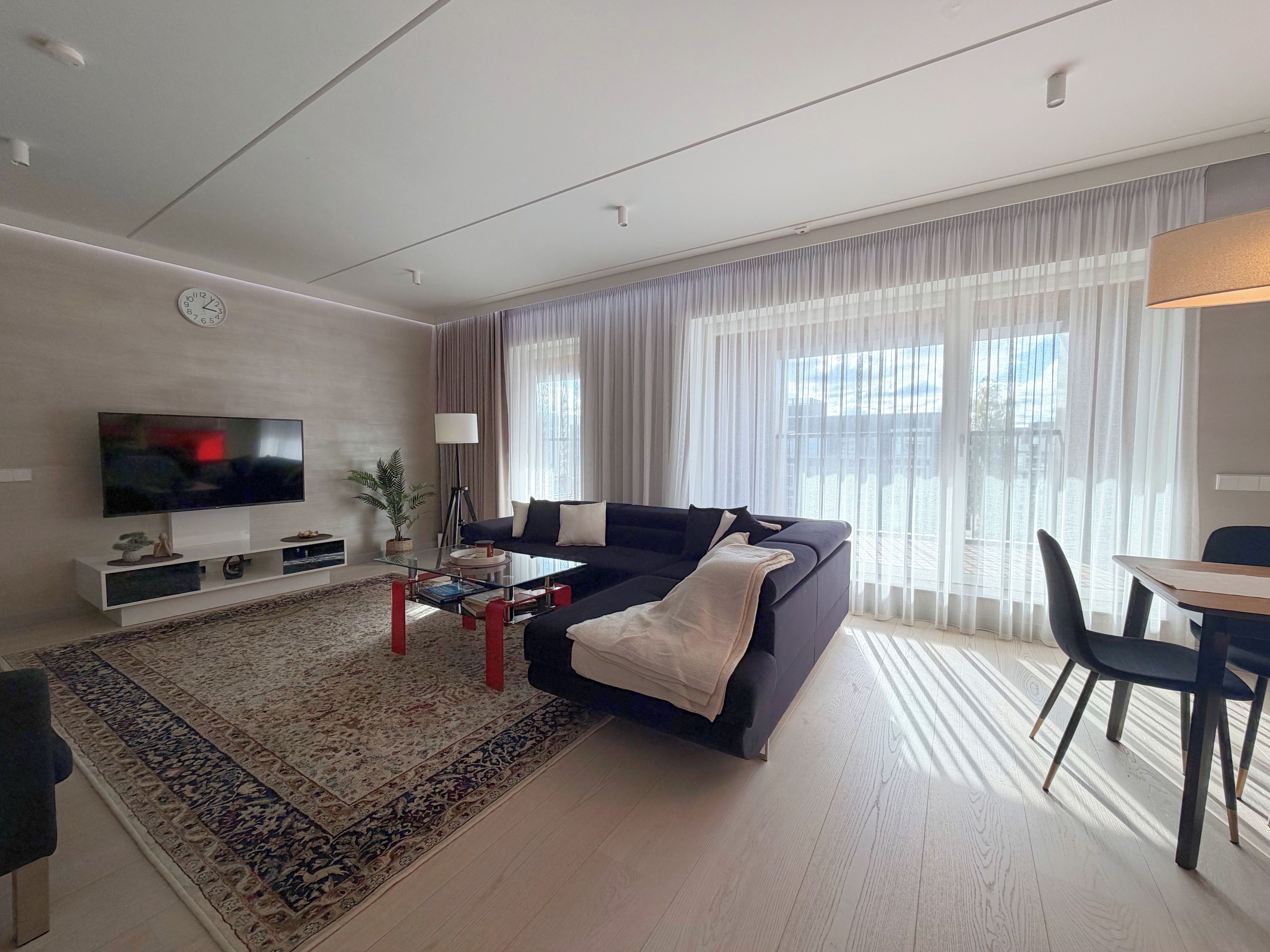 A modern living room with a flat-screen TV, a dark-colored sofa, a coffee table, and a dining area with a table and chairs.
