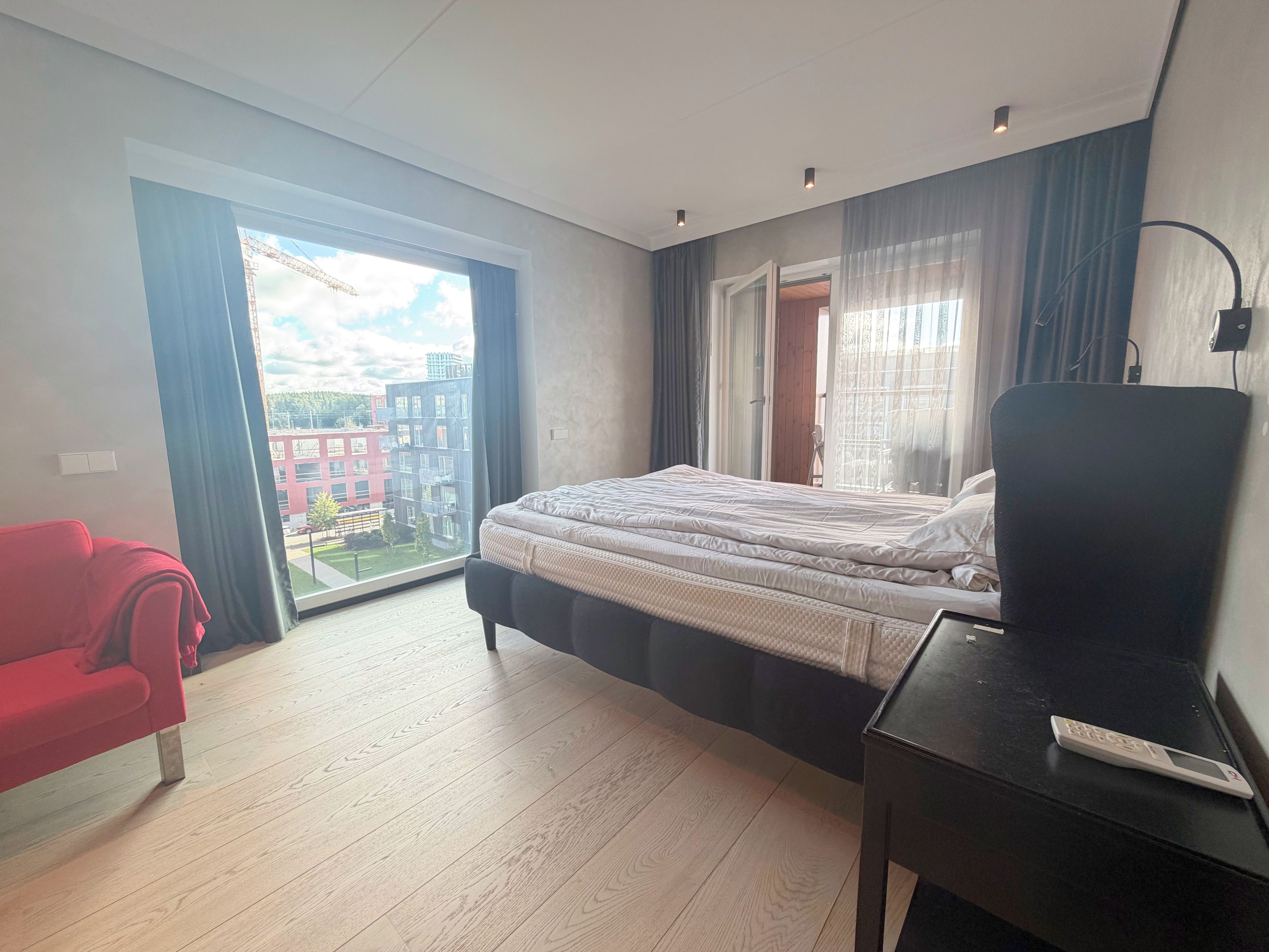 A modern bedroom with a large bed, a red chair, a desk, and a view of a building through a large window.