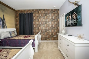 Ein Schlafzimmer mit Bücherregal, einem Bett mit gemustertem Überwurf, einem Schminktisch und einem an der Wand hängenden Bild.