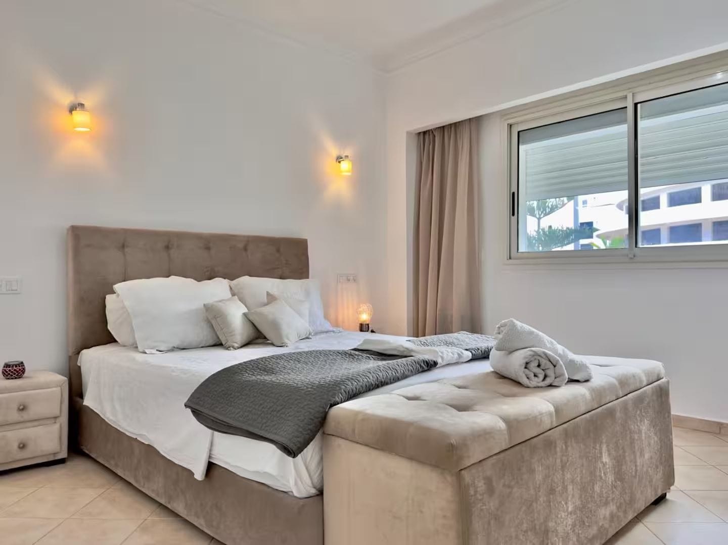 A bedroom with a bed, bedside tables, a window, and a view of buildings outside.